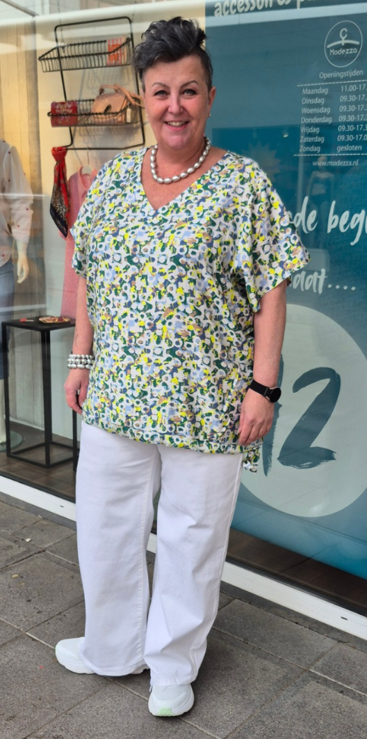Vrouw in grote maat draagt een vrolijke geprinte zomertop met witte broek voor de Modezza winkel in Veldhoven